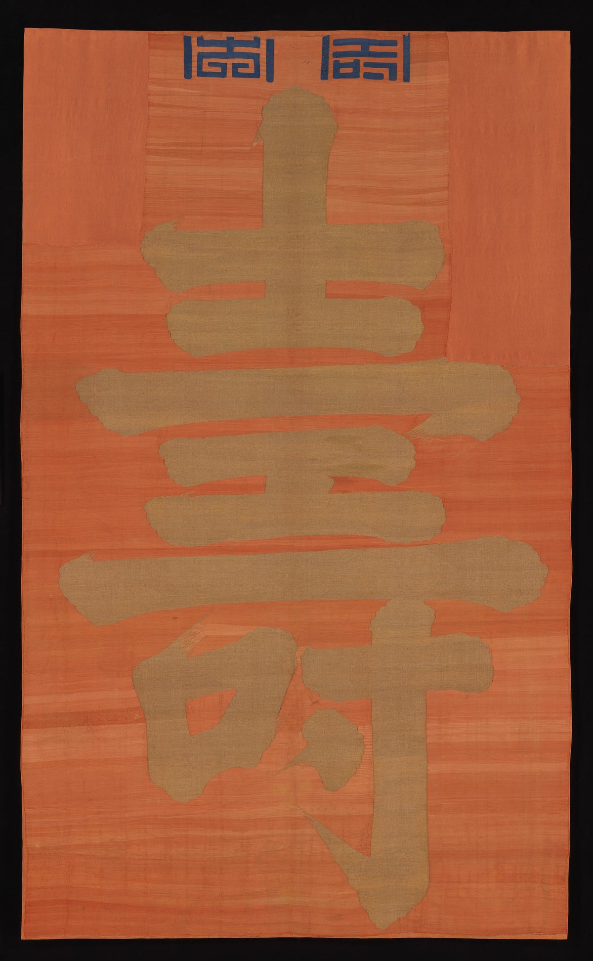 A large gold-colored shou character woven on an orange hanging, shown in full.